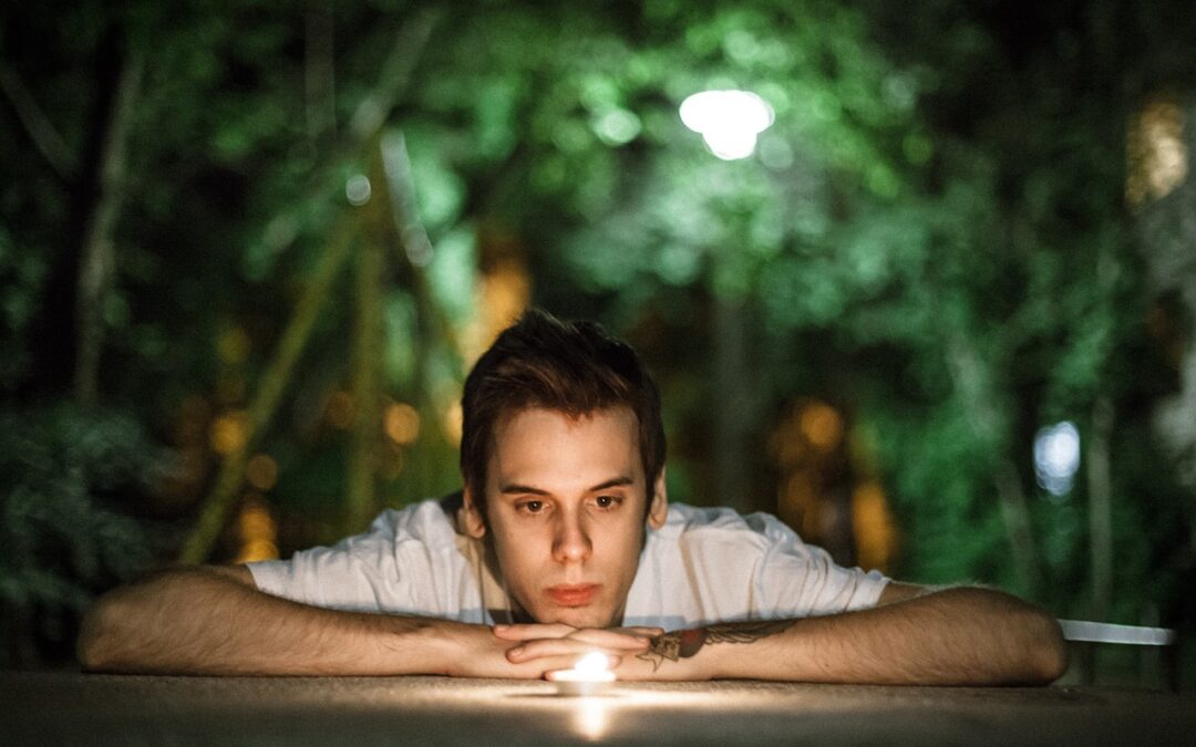 A man with short brown hair leans on a surface with his arms crossed, gazing thoughtfully at a small glowing light in front of him, set against a blurry, green outdoor background at night.