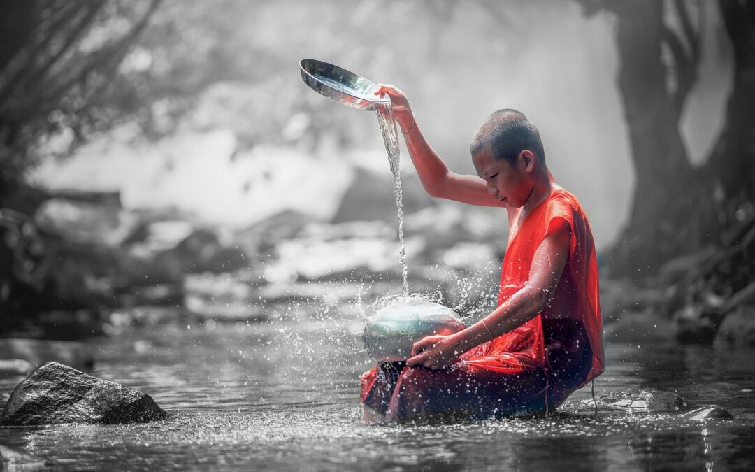 A young Buddhist monk in an orange robe kneels in a shallow stream, pouring water from a metal bowl onto another, surrounded by a misty, natural forest setting.