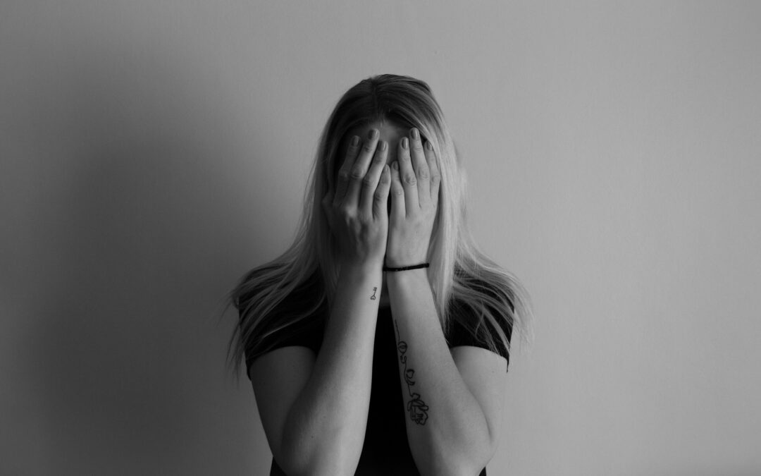 A person with long hair wearing a dark shirt stands against a plain background, covering their face with both hands. Visible tattoos and a hair tie are on their arms, and the image is in black and white.